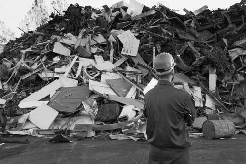 A man wearing a safety helmet and dark work clothing standing with his back to the camera, observing a large pile of mixed waste materials including broken furniture, plastic and metal debris, cardboard boxes, and electronic waste, all stacked on a paved surface in an outdoor setting. The waste appears haphazardly piled and consists of various textures such as smooth plastic, rough cardboard, and metallic surfaces. The scene may be associated with rubbish removal or clearance activities, typical of services provided by Garden Clearance Poplar in the local area. The background shows a natural environment with some trees and sky visible above the waste heap, suggesting an open space or yard used for waste collection or disposal. The lighting is natural, with a balanced exposure highlighting the contrasting textures and materials within the debris pile, indicative of a typical waste management site.
