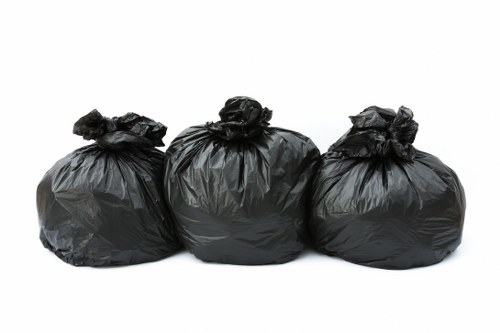 Three black plastic rubbish bags, tightly knotted at the top, are positioned in a row on a plain white surface. The bags are filled with waste materials, creating a rounded, bulky appearance with a slightly glossy finish, indicating they are made of standard bin liner material. Each bag exhibits irregular creases and folds, reflecting the uneven contents inside. The background is entirely white and unadorned, providing a clear focus on the bags without any environmental context, suggesting they are ready for collection by a local rubbish removal service such as Garden Clearance Poplar. The simplicity of the scene highlights the typical representation of waste awaiting disposal, relevant to garden clearance or general rubbish removal in a London suburb, possibly Poplar, as referenced on the associated webpage. The overall composition is clean, neutral, and professional, aligning with the context of waste management services dedicated to residential or commercial rubbish collection in the area.