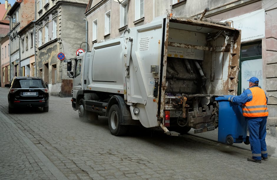 A narrow urban alleyway filled with a large pile of discarded cardboard boxes, plastic bags, and various waste materials stacked against a wall at the far end. In the foreground, a large, weathered sh