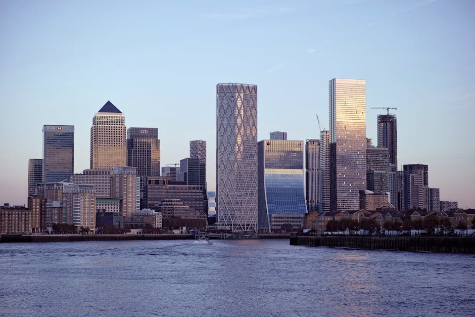 A nighttime cityscape featuring a skyline of modern office buildings illuminated with various coloured lights, including purple, red, and white, reflecting off the calm water in the foreground. The bu