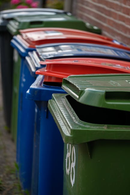 A row of large outdoor refuse bins positioned along a paved residential pathway next to a brick wall. The bins are made of durable plastic with textured surfaces on their removable lids. The bins are 